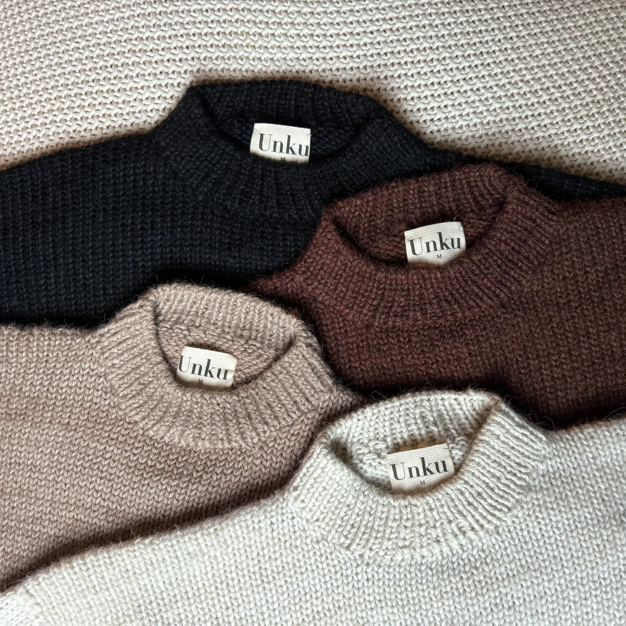 Four 100% peruvian baby alpaca sweaters in white, beige, brown, and black on a white textured background