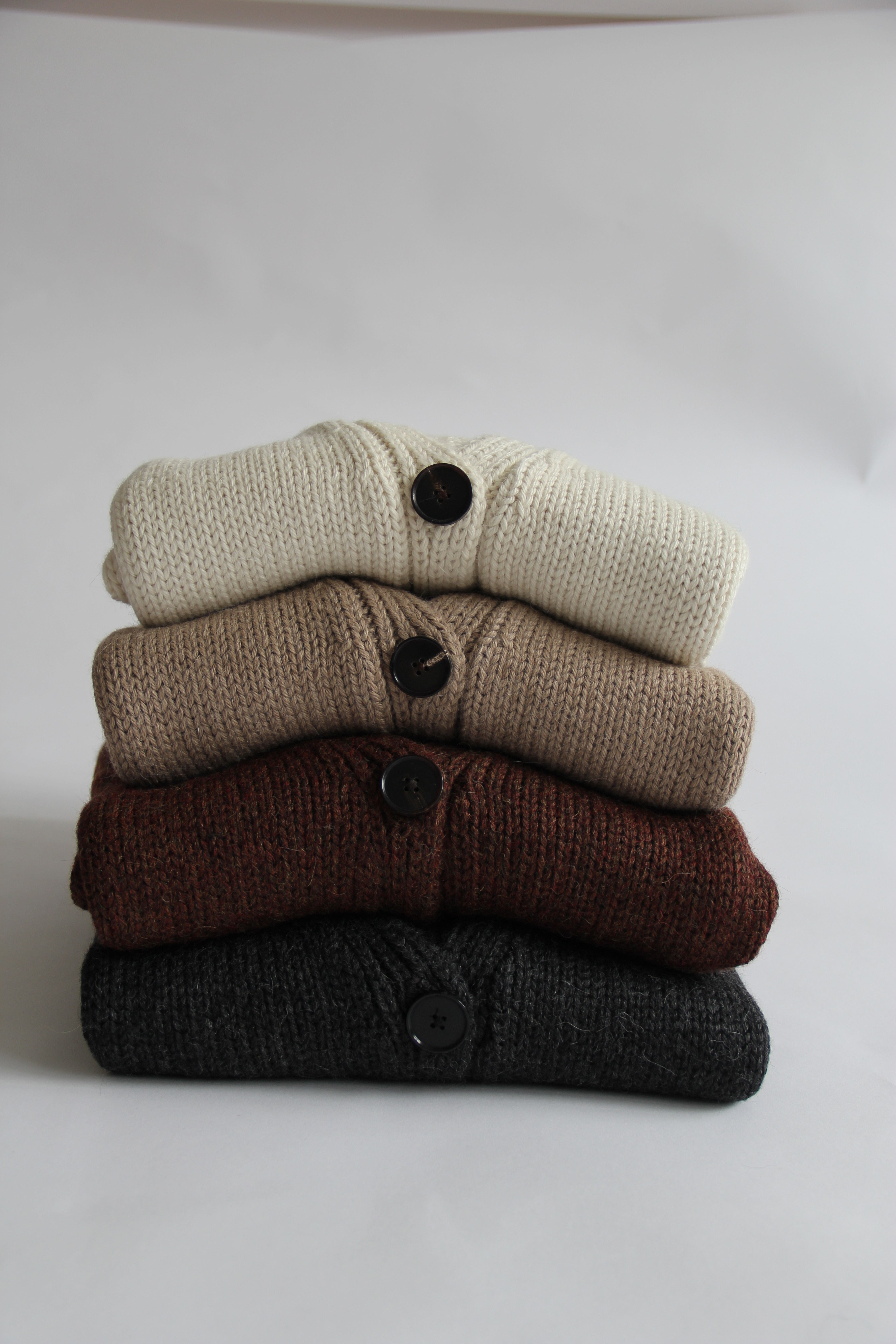Four rolled-up 100% Peruvian baby alpaca cardigans in white, beige, brown and charcoal on a white background.