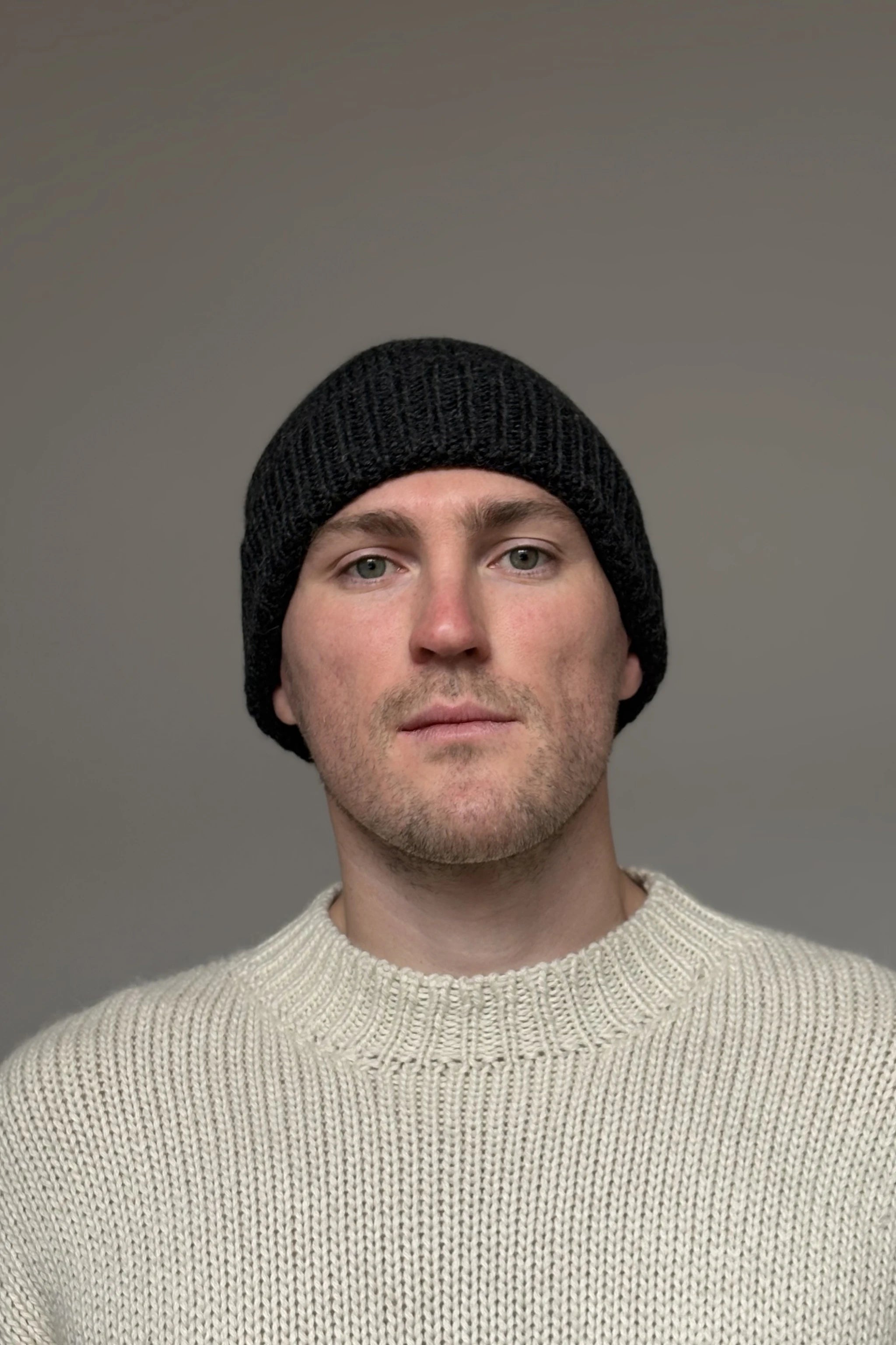 A man wearing a charcoal gray 100% Peruvian baby alpaca beanie in a white baby alpaca sweater with a neutral background 