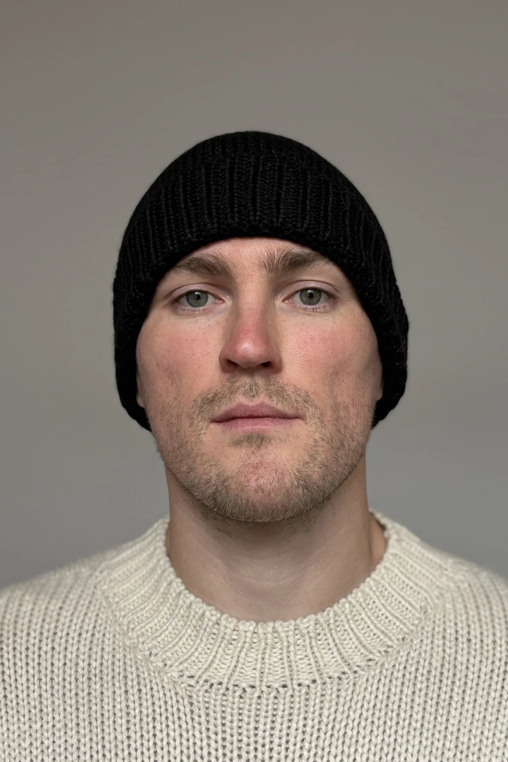 A man wearing a black 100% Peruvian baby alpaca beanie in a white baby alpaca sweater with a neutral background 