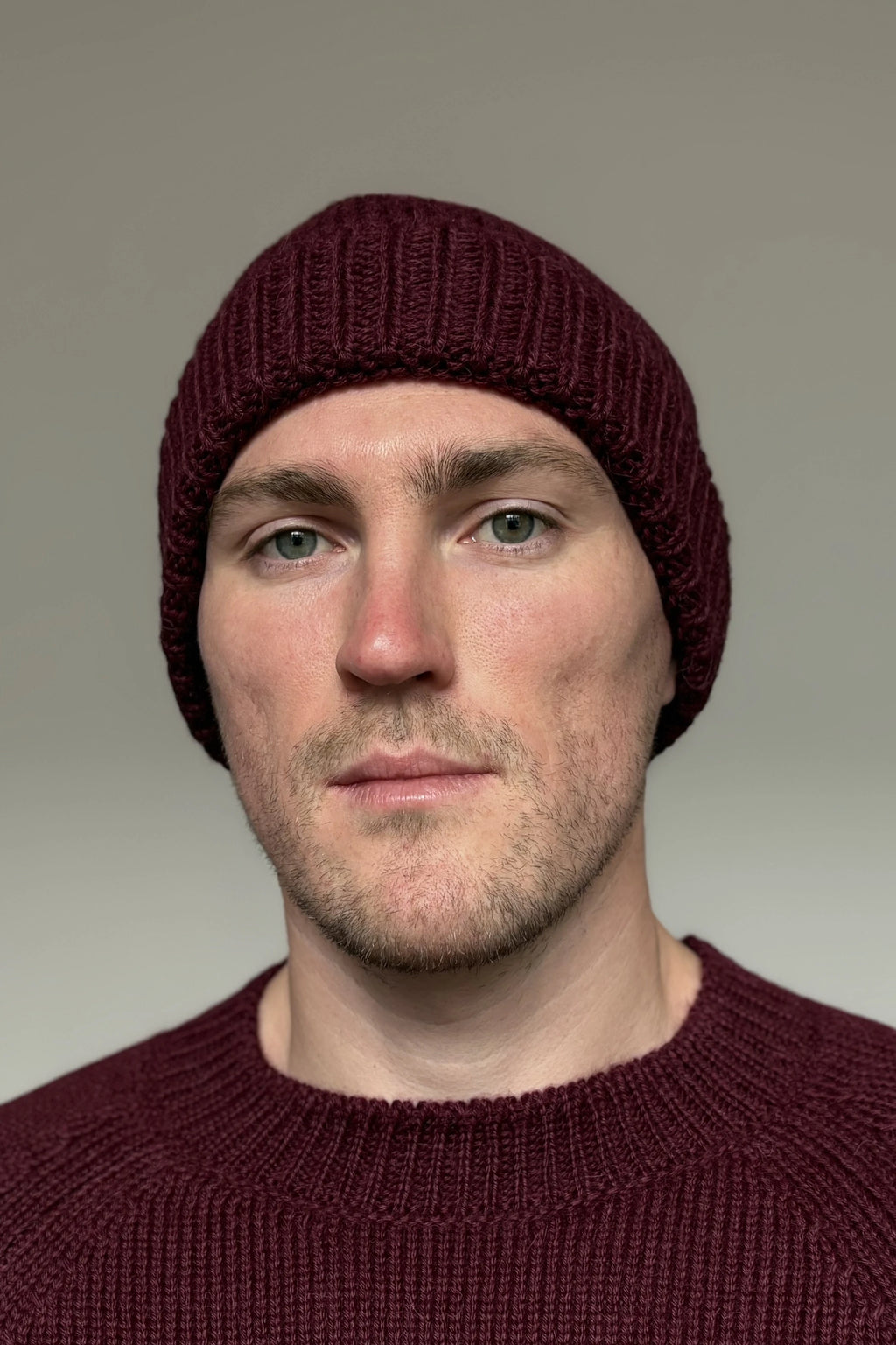 A man wearing a burgundy 100% Peruvian baby alpaca beanie and a burgundy baby alpaca sweater with a neutral background 