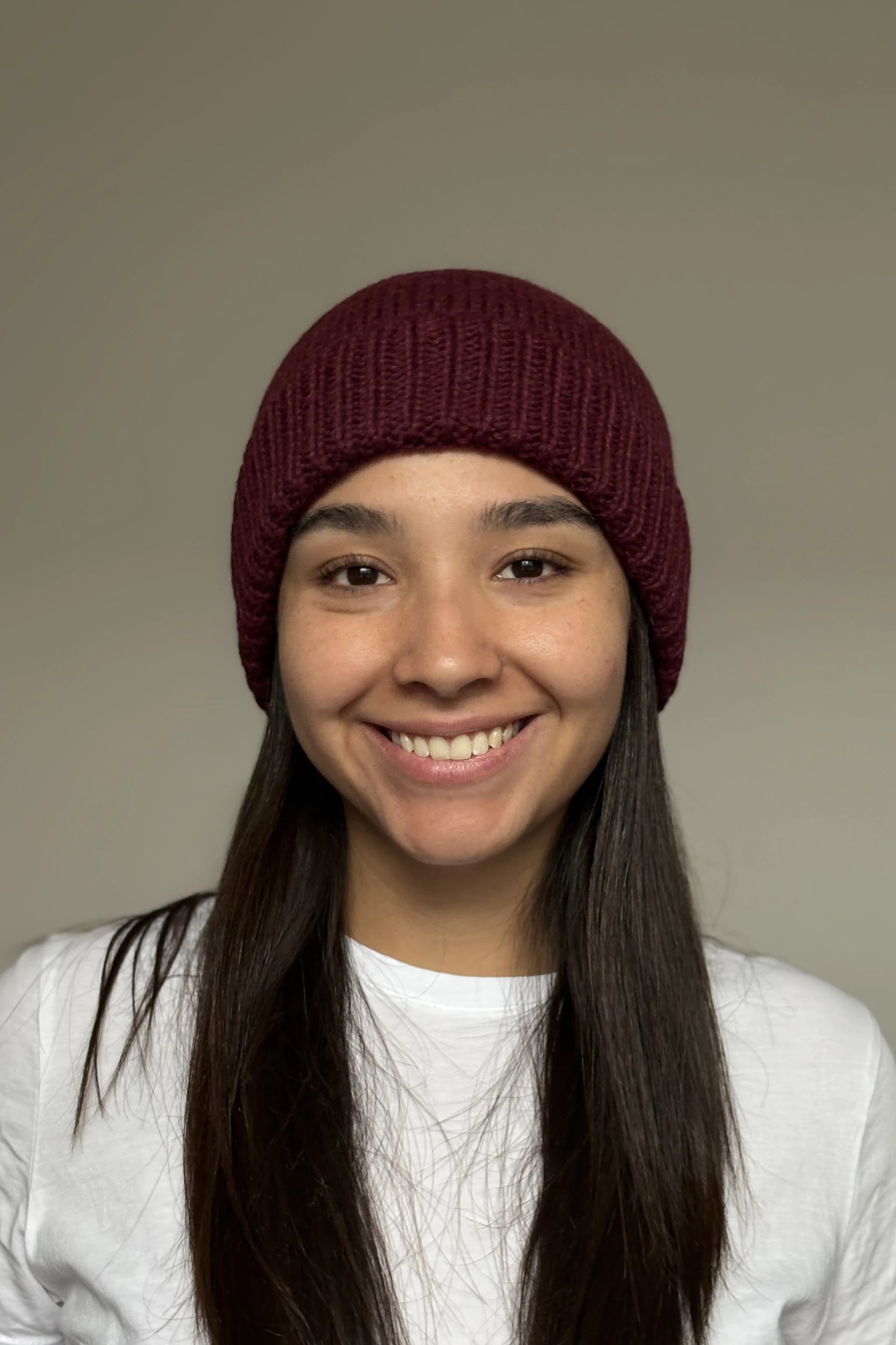 A beautiful young woman wearing a burgundy 100% Peruvian baby alpaca beanie in a white shirt with a neutral background 