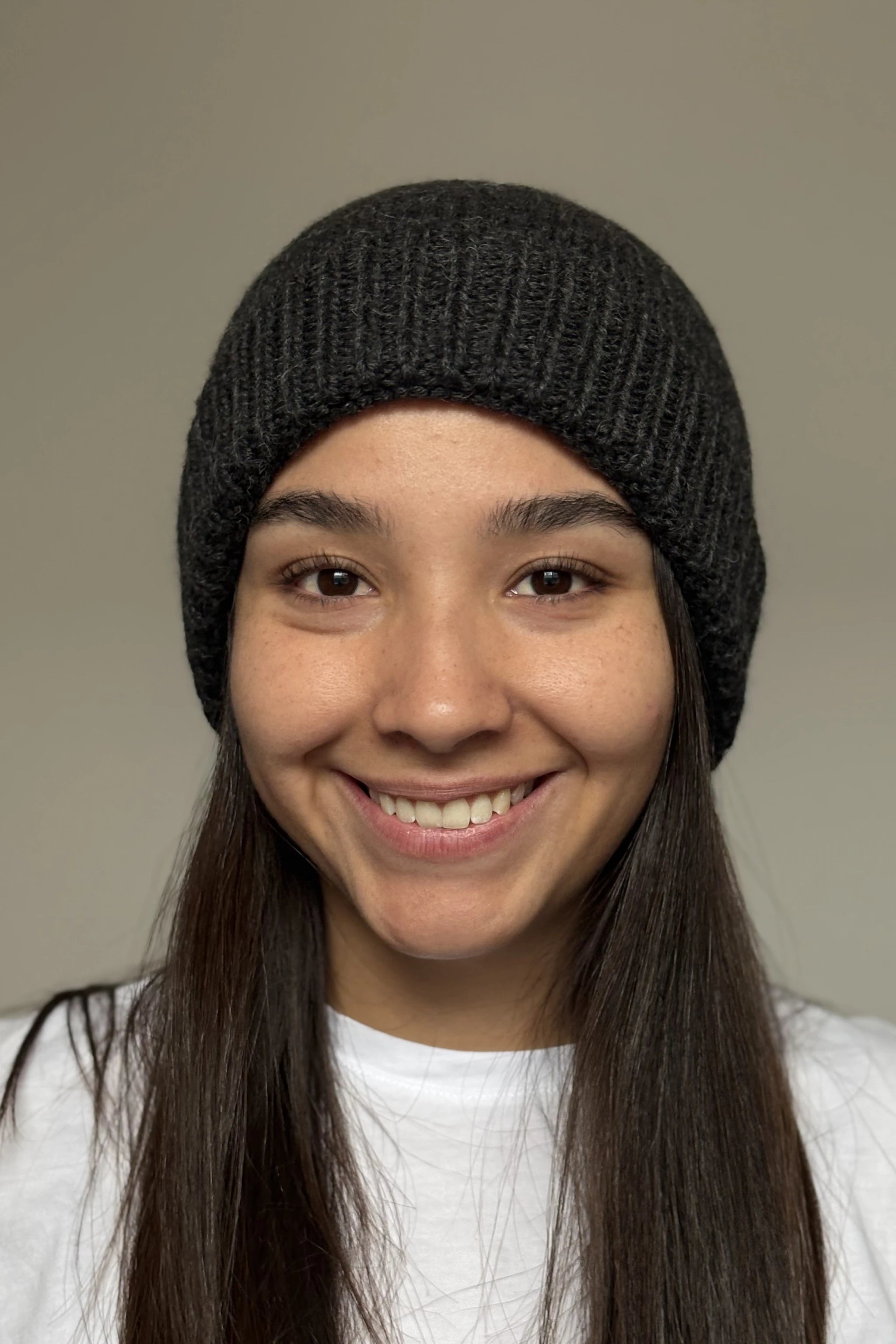 A beautiful young woman wearing a charcoal gray 100% Peruvian baby alpaca beanie in a white shirt with a neutral background 