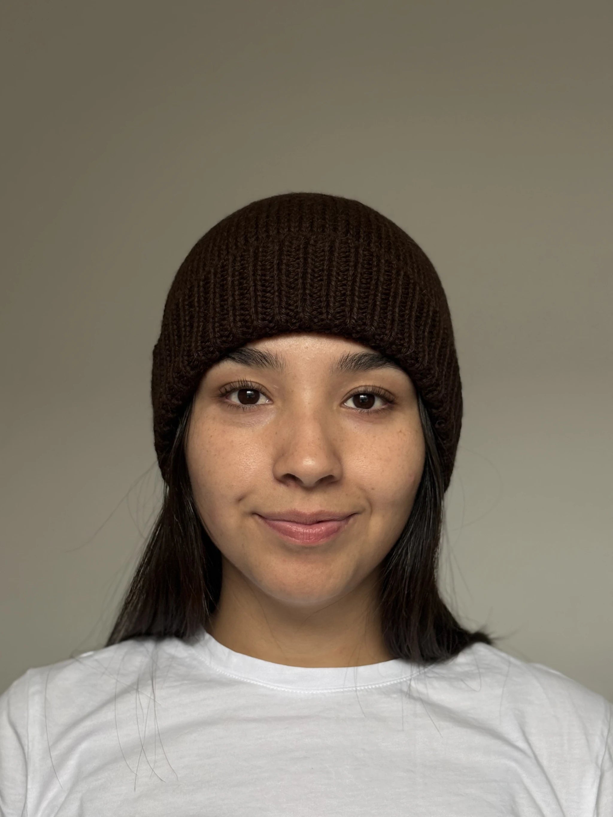 A beautiful young woman wearing a chocolate brown 100% Peruvian baby alpaca beanie in a white shirt with a neutral background 